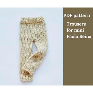 May include: Cream-coloured knitted trousers for a mini Paola Reina doll. The pants are handmade with a textured stitch and feature a small stain on the leg. The image includes text that reads "PDF pattern Trousers for mini Paola Reina."