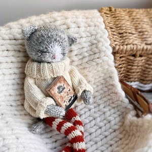May include: A gray knitted cat doll wearing a white sweater and red and white striped leggings. The doll is holding a small book with the text "Feliz Navidad" on the cover. The doll is sitting on a white knitted blanket.
