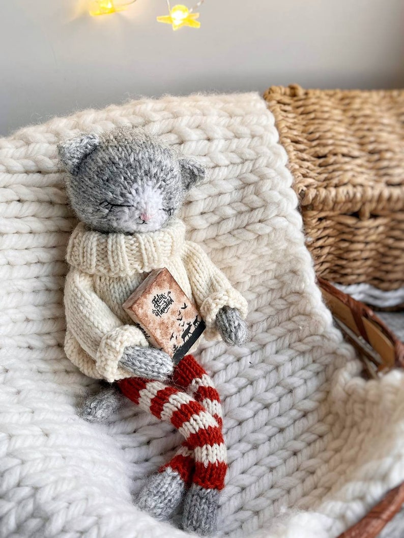 May include: A gray knitted cat doll wearing a white sweater and red and white striped leggings. The doll is holding a small book with the text "Feliz Navidad" on the cover. The doll is sitting on a white knitted blanket.