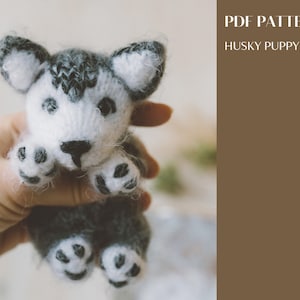 Könnte beinhalten: Ein gestricktes Husky-Welpenspielzeug mit schwarz-weißem Fell. Das Spielzeug wird in einer Hand gehalten. Das Bild enthält den Text "PDF PATTERN HUSKY PUPPY".