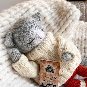 May include: A gray and white knitted cat toy wearing a cream sweater and red knitted pants is holding a small book that says "Feliz Navidad". The cat is lying on a white knitted blanket.