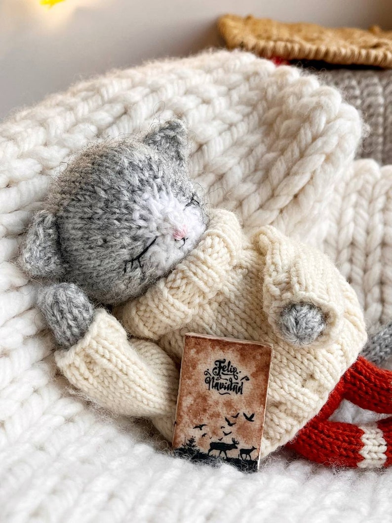 May include: A gray and white knitted cat toy wearing a cream sweater and red knitted pants is holding a small book that says "Feliz Navidad". The cat is lying on a white knitted blanket.