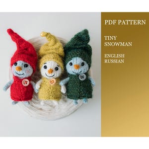 May include: Three knitted snowman ornaments in red, yellow, and green. The ornaments are sitting on a white ball of yarn. The text "PDF PATTERN TINY SNOWMAN ENGLISH RUSSIAN" is in the background.