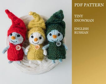 Puede incluir: Tres adornos de muñeco de nieve tejidos en rojo, amarillo y verde. Los adornos están sobre una bola de lana blanca. El texto "PDF PATTERN TINY SNOWMAN ENGLISH RUSSIAN" está en el fondo.