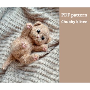 Pode incluir: Um gatinho de malha castanho claro com grandes olhos pretos e almofadas cor-de-rosa está deitado de costas sobre uma manta de malha cinzenta. Os braços do gatinho estão levantados. O texto à direita diz "PDF pattern Chubby kitten."
