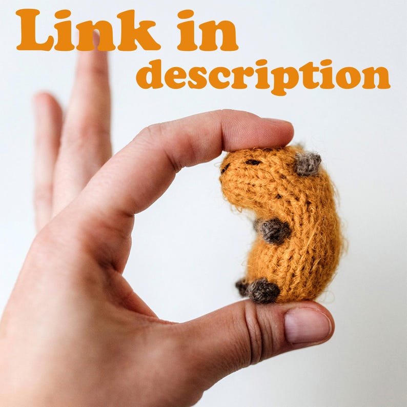 A tiny knitted capybara is held between fingers, showing its very small scale. The toy has a warm brown body with darker paws and a soft, fuzzy texture. The close-up highlights neat stitches and miniature proportions.