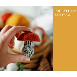 May include: A knitted mushroom toy with a red cap and a white and black striped stem. The mushroom is being held in a hand. The text "PDF PATTERN MUSHROOM" is in the background.