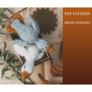 May include: Two knitted goslings, one blue and one white, with orange beaks, are lying on top of a book. The image is a close-up of the goslings, and the background is blurred. The text "PDF PATTERN MICRO GOSLING" is visible in the upper right corner of the image.
