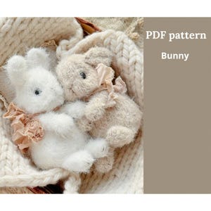 Pode incluir: Dois coelhos de pelúcia, um branco e outro bege, aninhados em um cobertor de malha creme. Cada coelho tem um pequeno laço franzido em volta do pescoço. A imagem inclui o texto "PDF pattern Bunny."