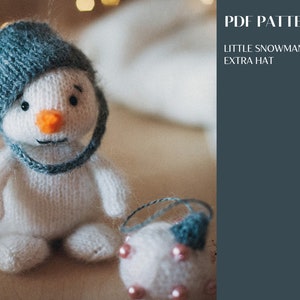 May include: A white knitted snowman toy with a blue hat, orange nose, and black eyes, accompanied by a small white ball with pink beads and a blue accent. The image includes text that reads PDF PATTERN LITTLE SNOWMAN + EXTRA HAT.