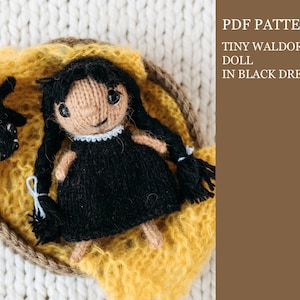 Puede incluir: Patrón PDF para una pequeña muñeca Waldorf con un vestido negro. La muñeca está tejida y tiene el pelo negro y los ojos marrones. La muñeca está tumbada en una cesta de punto amarilla.