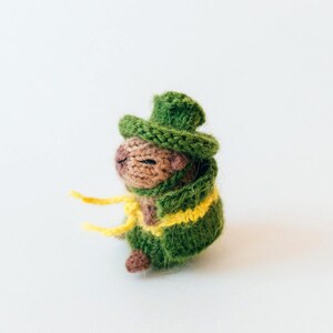 Side view of the knitted capybara wearing a green cape and matching hat. The yellow belt wraps around the body and hangs loosely in front. The photo shows the layered clothing and rounded body shape.