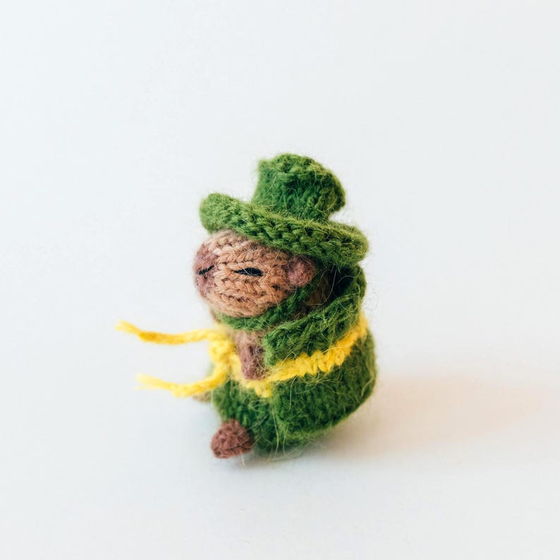 Side view of the knitted capybara wearing a green cape and matching hat. The yellow belt wraps around the body and hangs loosely in front. The photo shows the layered clothing and rounded body shape.