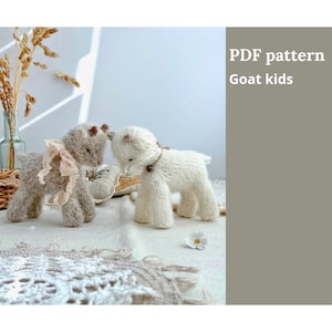 Peut inclure: Deux figurines de chèvre tricotées, une grise et une blanche, sont présentées sur une surface texturée. La chèvre grise a un ruban rose, tandis que la chèvre blanche a un collier marron. Le texte de l'image indique "PDF pattern Goat kids."