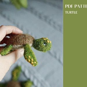 May include: A knitted turtle toy with a brown shell and green limbs. The turtle has yellow spots on its shell and green eyes. The toy is being held in a hand.