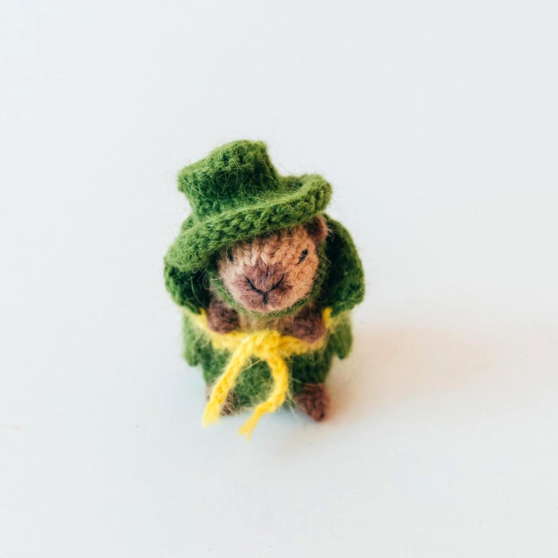 A miniature knitted capybara dressed in a green outfit stands upright on a white surface. The toy wears a small green hat and a yellow belt tied at the front. The soft yarn texture and tiny facial details are clearly visible.