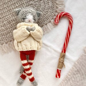 May include: A gray knitted cat doll wearing a white sweater and red and white striped pants. The doll is lying on a white surface with a candy cane next to it.