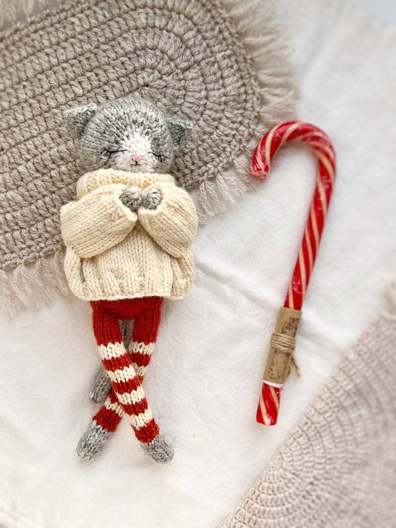 May include: A gray knitted cat doll wearing a white sweater and red and white striped pants. The doll is lying on a white surface with a candy cane next to it.