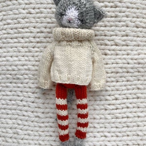 May include: A gray and white knitted cat doll wearing a cream colored sweater and red and white striped leggings.