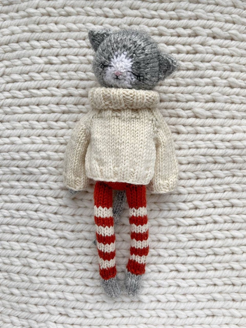 May include: A gray and white knitted cat doll wearing a cream colored sweater and red and white striped leggings.