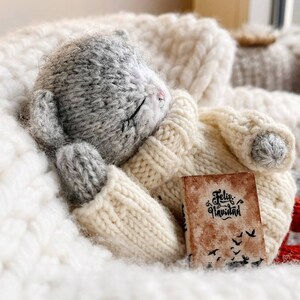 May include: A gray knitted cat toy wearing a white knitted sweater and holding a small book that says "Feliz Navidad". The cat is lying on a white fluffy blanket.