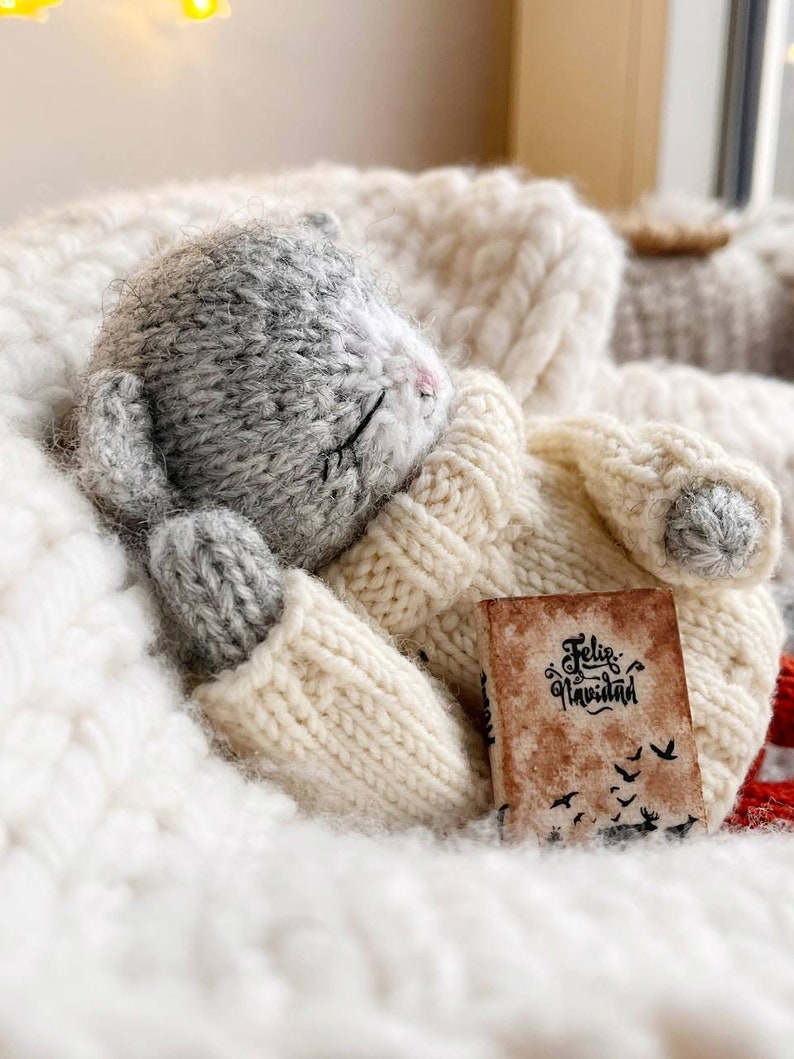 May include: A gray knitted cat toy wearing a white knitted sweater and holding a small book that says "Feliz Navidad". The cat is lying on a white fluffy blanket.