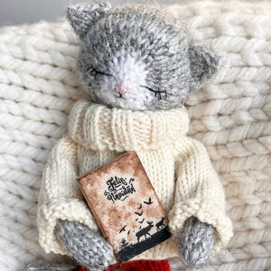 May include: A gray and white knitted cat doll wearing a white sweater and red and white striped pants. The doll is holding a small book with the text "Feliz Navidad" on the cover.