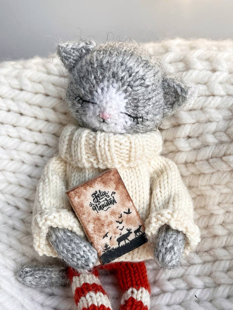 May include: A gray and white knitted cat doll wearing a white sweater and red and white striped pants. The doll is holding a small book with the text "Feliz Navidad" on the cover.