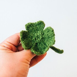 A tiny knitted shamrock is held in hand to show its scale. The shamrock is made in rich green yarn with clearly shaped leaves and a slim stem. The texture looks soft and slightly fluffy.