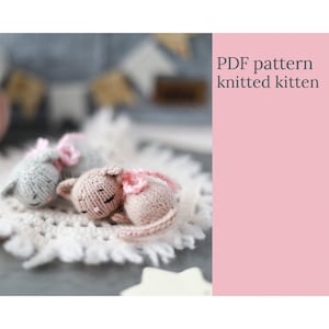 May include: Two knitted kitten toys, one gray and one pink, are curled up on a white, lacy surface. Each kitten has a pink scarf. The image includes the text "PDF pattern knitted kitten" on a pink background.
