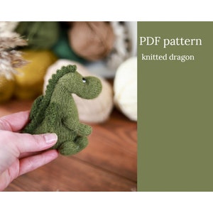 Könnte beinhalten: Ein kleiner, grüner, gestrickter Drachen mit strukturiertem Körper und stacheligem Rücken. Der Drache wird in einer Hand gehalten, mit dem Text "PDF pattern knitted dragon" im Hintergrund. Der Drache ist etwa 10 cm groß.