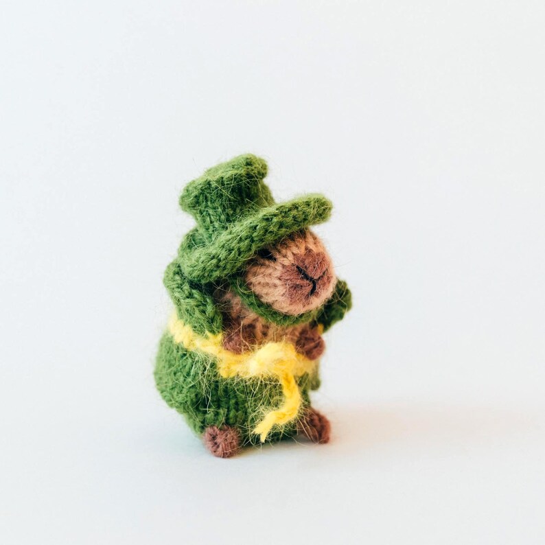 Angled view of the small knitted capybara in its St. Patrick outfit. The green hat sits slightly tilted, adding a playful look. The soft, slightly fuzzy yarn gives the toy a cozy handmade feel.