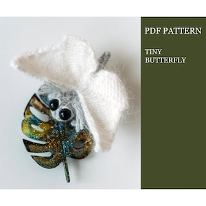 May include: A white crocheted butterfly with black eyes sits on a gold and blue glitter leaf. The text "PDF PATTERN TINY BUTTERFLY" is in the top right corner of the image.