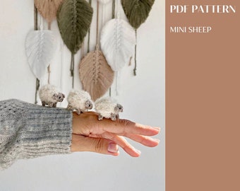 May include: A hand holding three small crocheted sheep. The sheep are white with gray faces and are standing in a row on the hand. The background is a wall with macrame wall hangings.