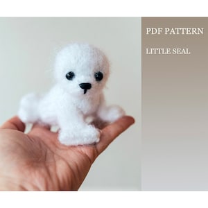 May include: A white crocheted seal amigurumi toy with black eyes and a black nose. The toy is sitting in a person's hand. PDF PATTERN LITTLE SEAL.