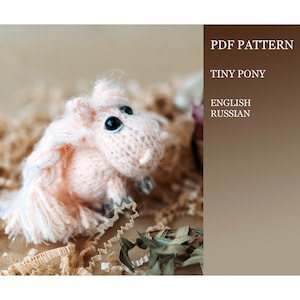 May include: A pink knitted tiny pony toy with a white mane and tail. The toy is sitting on a brown paper surface. PDF pattern for knitting a tiny pony in English and Russian.