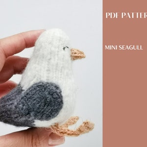 Może przedstawiać: Pluszowa zabawka amigurumi w kształcie mewy, wykonana z białej i szarej przędzy, z brązowym dziobem i brązowymi nogami z przędzy. Zabawka jest trzymana w dłoni. W tle widoczny jest tekst "PDF PATTERN MINI SEAGULL".