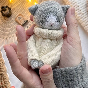 May include: A small gray knitted cat wearing a white knitted sweater. The cat is being held in a person's hands.