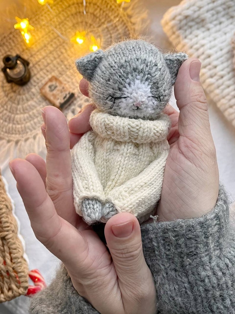 May include: A small gray knitted cat wearing a white knitted sweater. The cat is being held in a person's hands.