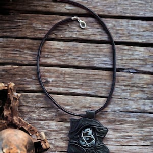 May include: A black leather necklace with a pendant featuring a dark grey stone with white veins. The pendant is shaped like a flower with intricate details. The necklace is photographed on a wooden surface.