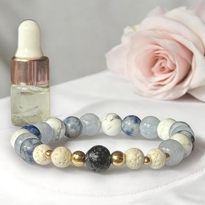 Puede incluir: Un brazalete de cuentas azules, blancas y doradas. Las cuentas tienen diferentes texturas y patrones. Una pequeña botella de vidrio con un cuentagotas está al lado del brazalete. También hay una rosa rosa.