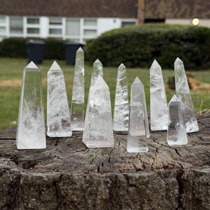 May include: A collection of clear quartz crystal obelisks of varying sizes, standing upright on a weathered wooden surface. The crystals are transparent with internal fractures, reflecting light. The background shows a grassy area and a building.