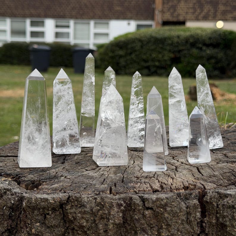 May include: A collection of clear quartz crystal obelisks of varying sizes, standing upright on a weathered wooden surface. The crystals are transparent with internal fractures, reflecting light. The background shows a grassy area and a building.