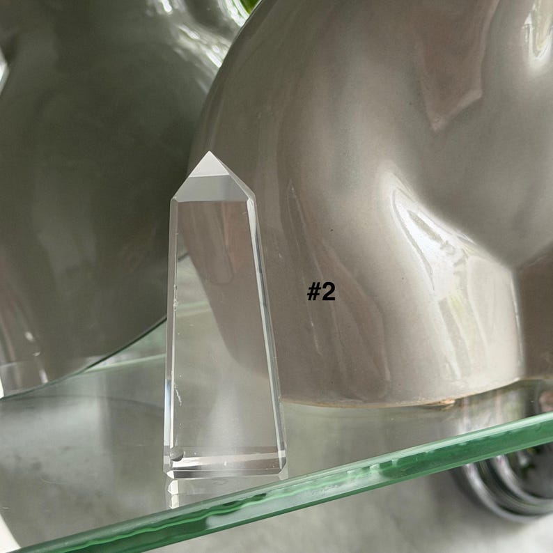May include: A clear crystal obelisk with a pointed top, standing upright. The crystal is transparent, allowing light to pass through. The number "#2" is printed on the side. The obelisk is displayed on a glass shelf.