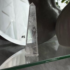 May include: Clear quartz obelisk with a pointed top. The crystal is transparent with internal inclusions and the text "#5" is visible on the side. The obelisk is displayed on a glass shelf.