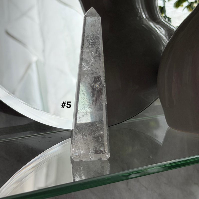 May include: Clear quartz obelisk with a pointed top. The crystal is transparent with internal inclusions and the text "#5" is visible on the side. The obelisk is displayed on a glass shelf.