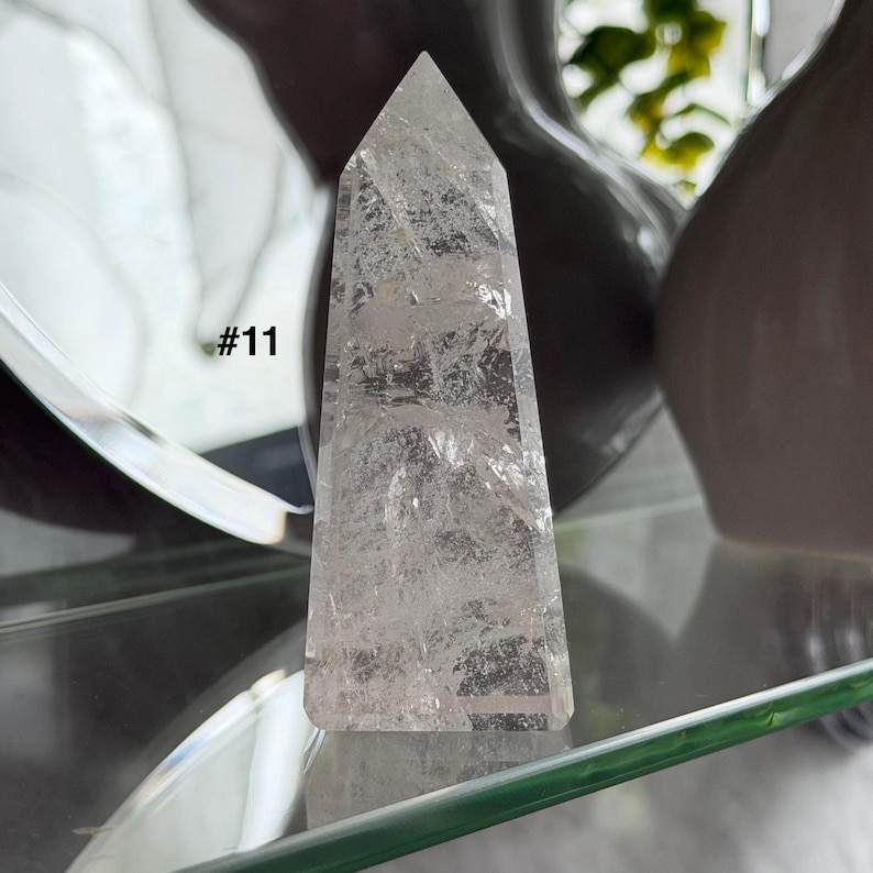 May include: A clear quartz crystal obelisk with a pointed top. The crystal has internal fractures and inclusions, giving it a textured appearance. The number "#11" is visible on the left side of the crystal. The obelisk is displayed on a glass shelf.