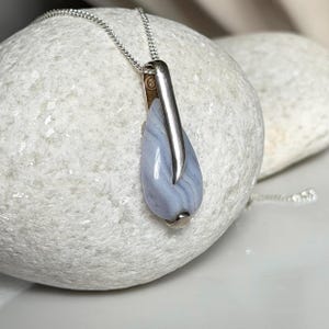 May include: A silver necklace with a teardrop-shaped pendant. The pendant features a light blue stone with a silver metal accent. The necklace is displayed on a white stone.
