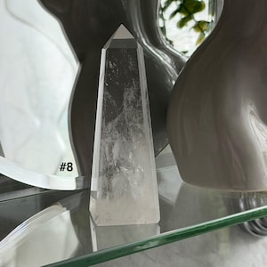 May include: A clear quartz crystal obelisk with a pointed top. The crystal is displayed on a glass shelf, with the number "#8" visible in the background. The obelisk is a light, transparent color.