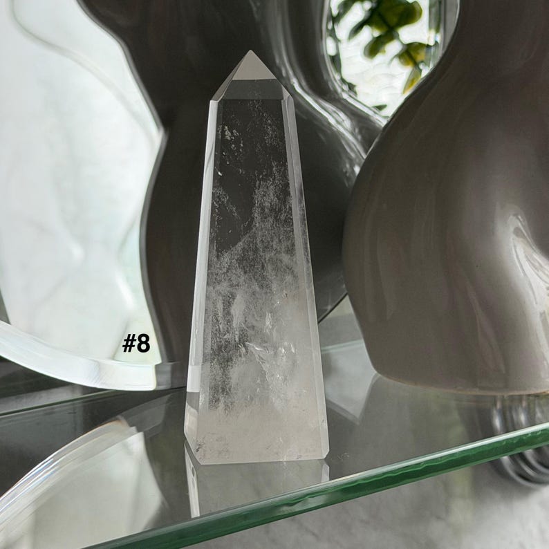 May include: A clear quartz crystal obelisk with a pointed top. The crystal is displayed on a glass shelf, with the number "#8" visible in the background. The obelisk is a light, transparent color.